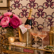 Load image into Gallery viewer, Close-up of Reese Way wallpaper by Pickwick &amp; Elm behind a brass bar cart with champagne, crystal glassware, pink peonies, and cocktail accessories.
